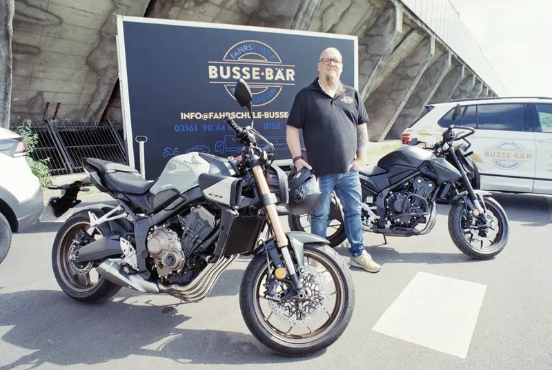 Michael Busse mit zwei Fahrschulmotorrädern vor dem Werbeanhänger der Fahrschule